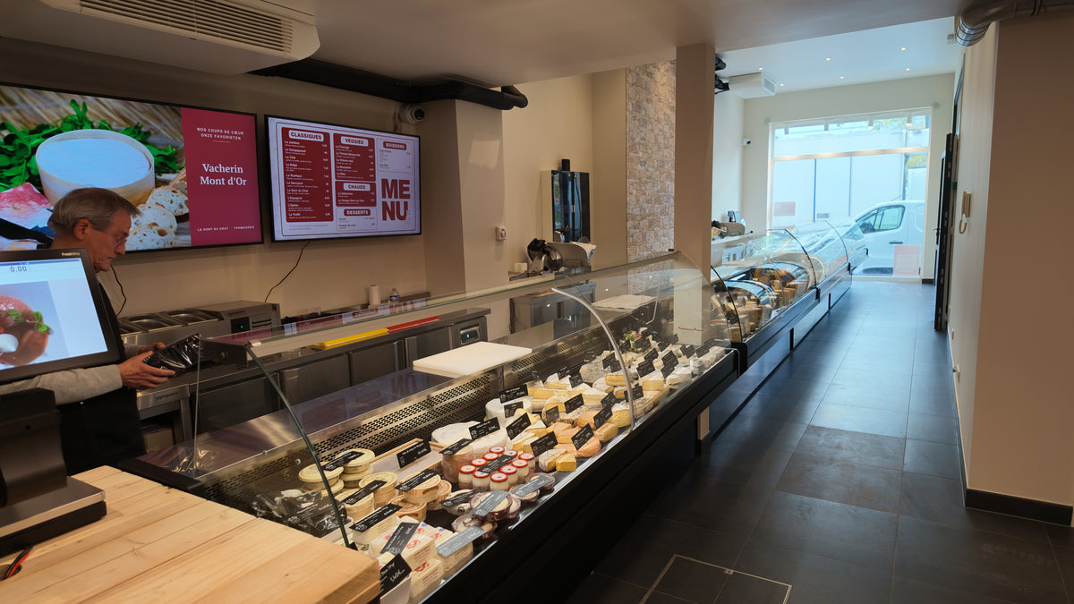 Intérieur de la boutique La Dent du Chat Place du Châtelain à Ixelles : comptoir à fromages et écrans du menu sandwichs
